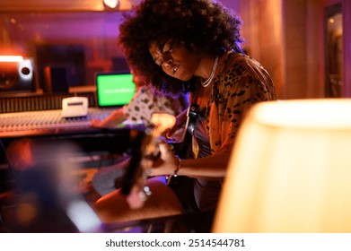 Professional talented multicultural female bass player sitting at music recording studio with bass guitar and recording music. There is a sound technician in a blurry background with channel mixer. - Powered by Shutterstock