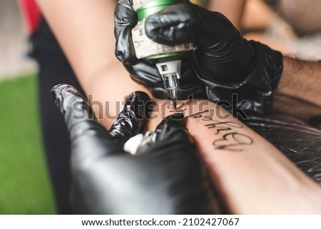 A professional tattoo artist uses a rotary tattoo machine with a round liner cartridge to draw cursive names on a female client's forearm.