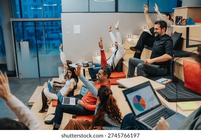 Professional team participates in a collaborative workshop, raising hands and engaging with laptops and charts. A dynamic office setting shows group learning and teamwork.