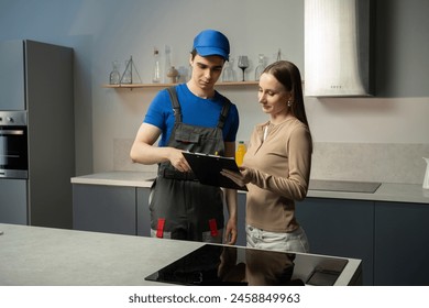 A professional technician in a blue cap and work uniform is showing a document or digital device to a female homeowner in a contemporary kitchen setting. They are engaged in a discussion, possibly - Powered by Shutterstock