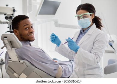 Professional Teeth Cleaning. Black Female Dentist Having Treatment With Male Patient, Professional Stomatologist Woman In Medical Mask And Face Shield Holding Dental Tools And Looking At Camera