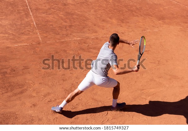 Professional tennis player\
performs forehand hit on clay tennis court. Young male athlete with\
tennis racket in action. Junior tennis sport. Back view, shadow,\
copy space