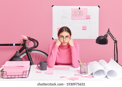 Professional tired female architect works on building sketch feels tired of long hours working checks graphic outlay tries to improve idea of planning poses at desktop surrounded with papers stickers - Powered by Shutterstock
