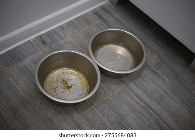 A professional top-down shot of two silver metal pet bowls sitting on a grey wood-textured tile floor. One bowl shows dried food residue and stains, while the other contains a small amount of water, captured in clear, neutral indoor lighting.