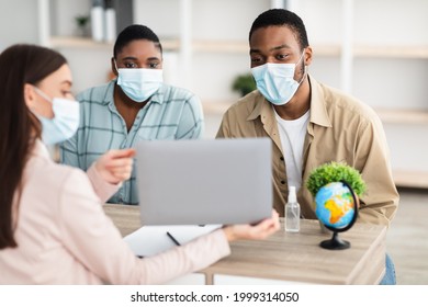 Professional Tour Agent Consulting Customers Couple Showing Laptop Recommending Vacation Destination For Black Spouses Sitting In Travel Agency Office. Selective Focus On Toursits Wearing Face Masks