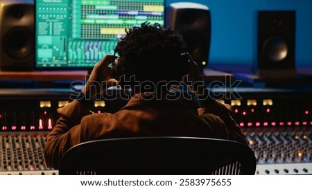 Professional tracking engineer using headphones to edit recorded tracks in control room, motorized sound control knobs. Audio expert working on mixing console and editing software. Camera A.