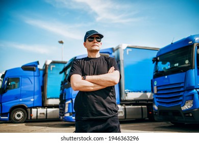 Professional Truck Driver With Hat And Sunglasses Confidently Standing In Front Of Big And Modern Truck Blue Truck Fleet. Bright Sunny Day. People And Transportation Concept.