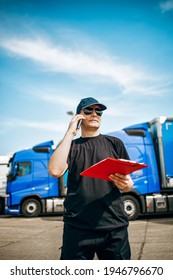 Professional Truck Driver With Hat And Sunglasses Confidently Standing In Front Of Big And Modern Truck Blue Truck Fleet. Bright Sunny Day. People And Transportation Concept.