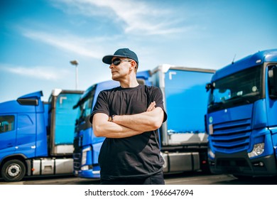 Professional Truck Driver With Hat And Sunglasses Confidently Standing In Front Of Big And Modern Truck Blue Truck Fleet. Bright Sunny Day. People And Transportation Concept.