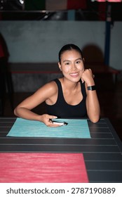 A professional vertical medium shot of a happy young Asian Thai woman sitting at an outdoor dining table at night. She is looking directly into the camera lens with a warm, cheerful smile and steady eye contact. Her left elbow rests on the dark table surface while her chin lightly leans against closed fist, displaying a smartwatch on wrist. In right hand, she casually holds a modern smartphone over a textured turquoise placemat. She is dressed in a simple black tank top with her dark hair neatly tied back. The background shows a dimly lit evening restaurant or cafe patio setting with a soft, out-of-focus background.