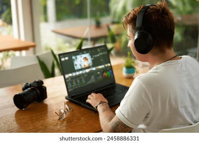 Professional video editor works on laptop in cozy cafe. Freelancer with tattoos wearing headphones edits film, digital camera on desk surrounded by plants by large window, daylight creative process. - Powered by Shutterstock