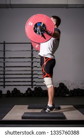 Professional weightlifter in knee wraps performs an exercise Snatch with the weights of 70 kilograms on the weightlifting platform with the barbell bars and dumbbells on the background.