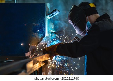 Professional Welder Wearing Protective Mask Performs Work At Steel Structure Manufacturing Plant. Yellow Sparks Spill Around.