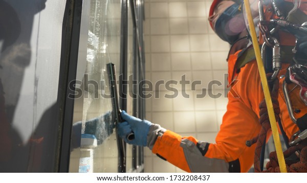 professional window cleaner work industrial\
climber in helmet and gloves holds a screed on a dirty window\
close-up in sunny weather at high\
altitude