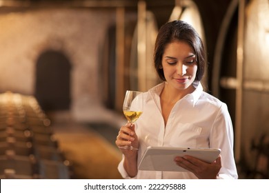 Professional winemaker female tasting a glass of white wine in his traditional cellar surrounded by wooden barrels. She uses a digital tablet - Powered by Shutterstock