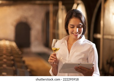 Professional winemaker female tasting a glass of white wine in his traditional cellar surrounded by wooden barrels. She uses a digital tablet - Powered by Shutterstock