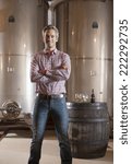 Professional winemaker looking at camera and tasting a glass of white wine in his cellar with modern stainless steel drums and traditional wooden barrels