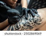 professional woman artist making stylish black tattoo of Japanese demon mask on man hand with machine and needle in salon, shoulder body art