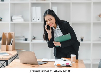 professional woman in black suit is engaged in phone conversation while reviewing documents on her laptop. She appears focused and productive in modern office setting. - Powered by Shutterstock