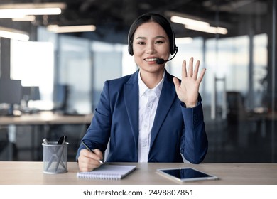 Professional woman in a business suit wearing a headset, looking at the camera, and waving while talking during a video call in a modern office setting. - Powered by Shutterstock