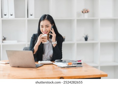 professional woman is engaged in phone conversation while enjoying cup of coffee at her desk. She is focused on her laptop, surrounded by notebooks and stationery, creating productive atmosphere. - Powered by Shutterstock