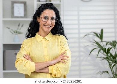 A professional woman with glasses stands confidently, her arms crossed, in a well-lit office environment, conveying empowerment and positivity. - Powered by Shutterstock