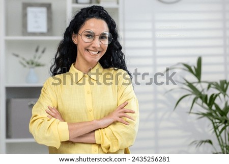 A professional woman with glasses stands confidently, her arms crossed, in a well-lit office environment, conveying empowerment and positivity.