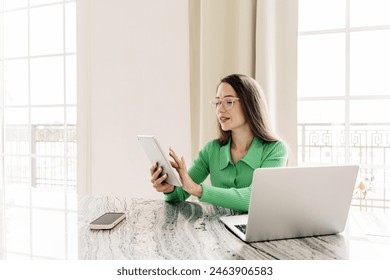 A professional woman with glasses, wearing a green sweater, using a tablet while working at a laptop.

 - Powered by Shutterstock