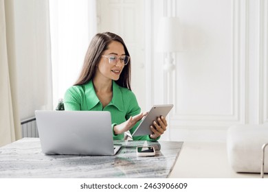 A professional woman with glasses, wearing a green sweater, using a tablet while working on a laptop.

 - Powered by Shutterstock