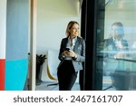 Professional woman in a gray blazer checks her smartphone while standing near a window reflecting her image in a contemporary office