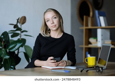 Professional woman holding a smartphone and looking thoughtful at her desk with a laptop, coffee mug, and notebook in a modern office. - Powered by Shutterstock