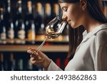 Professional woman sommelier smells white wine from glass, standing in cellar against shelf various alcoholic beverage background.