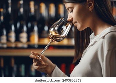 Professional woman sommelier smells white wine from glass, standing in cellar against shelf various alcoholic beverage background. - Powered by Shutterstock