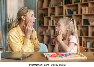 Professional woman speech therapist studying together with pretty little girl, learning practice pronunciation exercises with child at office, touching faces and grimacing, sitting at desk - Shutterstock ID 2198746363