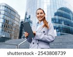 A Professional Woman Using a Tablet Outside Modern Office Buildings on a Bright Day in the City