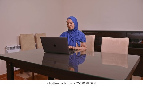 Professional woman wearing a blue hijab focused on her laptop while working at a desk in an office setting. - Powered by Shutterstock