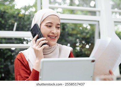 A professional woman wearing braces and a hijab is smiling while multitasking through a phone call and document review, showcasing a modern and organized lifestyle. - Powered by Shutterstock