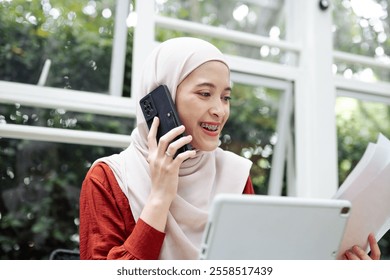 A professional woman wearing braces and a hijab is smiling while multitasking through a phone call and document review, showcasing a modern and organized lifestyle. - Powered by Shutterstock