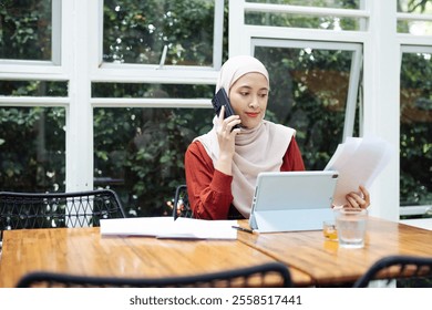 A professional woman wearing braces and a hijab is smiling while multitasking through a phone call and document review, showcasing a modern and organized lifestyle. - Powered by Shutterstock