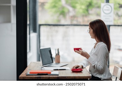 Professional woman in white shirt working on a laptop at a wooden desk, holding a red coffee cup, in a bright and modern office environment. - Powered by Shutterstock