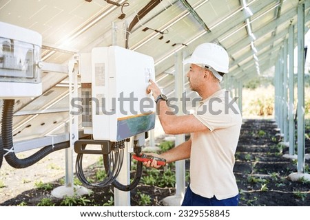 Professional worker checking voltage on solar inverter. Male using current probe to measure output voltage. Man in helmet pointing at inveter dial on back side of PV panel.