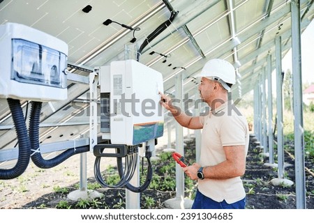 Professional worker checking voltage on solar inverter. Male using current probe to measure output voltage. Man in helmet pointing at inveter dial on back side of PV panel.