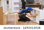 Professional worker in overalls consults furniture assembly instructions from laptop. Worker wearing a cap.