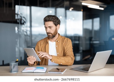 Professional worker using a digital tablet while sitting at an office desk. The workspace features a laptop, notepads, and modern decor, signifying digital communication