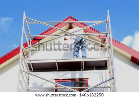 Professional workman is painting exterior walls and wooden window frames of ancient house at scaffold tower, outside home renovation in close up under sunny blue sky