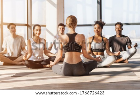 Professional Yoga Coach Having Lesson With Group Of Young People, Sitting In Lotus Position Indoors Together, Rear View Of Trainer
