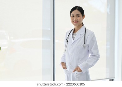 Professional young Asian female doctor in uniform, standing in the hospital, hands in pocket, smiling and looking at camera. - Shutterstock ID 2223600105