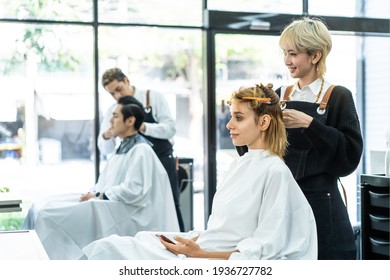 Professional Young Asian Female Hairdresser Or Hair Stylist Talking, Laughing, Using Scissors To Cut Beautiful Caucasian Woman Customer's Blonde Hair Sitting With Confidence In A Modern Beauty Salon.