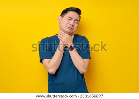 Professional young Asian male doctor or nurse wearing a blue uniform feeling sick and having neck pain, suffering from discomfort after long hours of sedentary isolated on yellow background