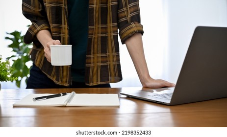 Professional Young Asian Male Freelancer Or Programmer In Flannel Shirt Leaning On His Office Desk, Holding A Mug Of Coffee. Cropped Image